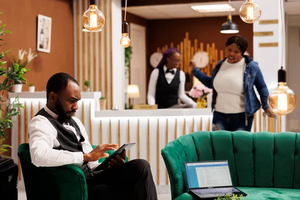 アフリカ系アメリカ人男性のホテル経営者は デジタルタブレット管理予約を使用してロビーに制服を着て ベルボーイは 左荷物レジスタに詳細を入力して電子機器を保持 — ストック写真