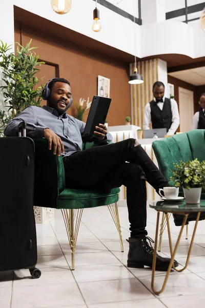 快乐的年轻非洲裔美国人坐在宾馆休息区 一边用数字平板电脑和无线耳机 一边微笑着看着摄像头 旅游时用录像与家人交谈 — 图库照片