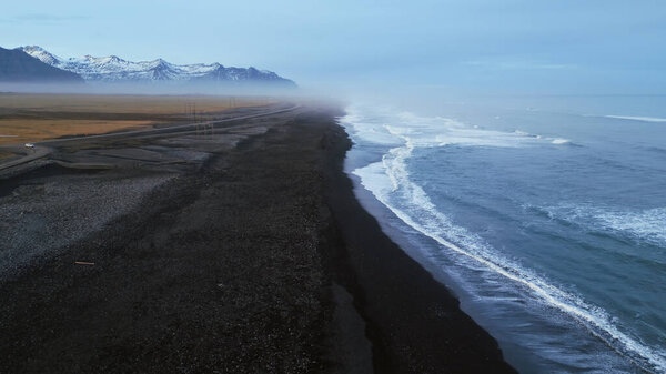 Исландский черный песчаный пляж дрон выстрел, красивая береговая линия с волнами, разбивающимися на атлантическом берегу. Северный ландшафт с заснеженными горами и черным песком на пляжах, живописный маршрут. Медленное движение.