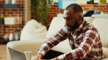 Odaklanmış Afro-Amerikan evde kalıp bir fincan kahvenin tadını çıkarırken dizüstü bilgisayardaki bilgilere bakıyor. Modern konforlu apartman dairesinde uzaktan tamamlayan tele-işçi çalışanı