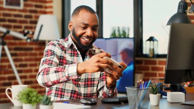 Akıllı telefondan video oyunu oynayıp kendini eğlendiren neşeli, tutkulu bir oyuncu. Gülümseyen rekabetçi Afrikalı Amerikalı adam hoş bir apartman dairesinde telefonda eğleniyor.