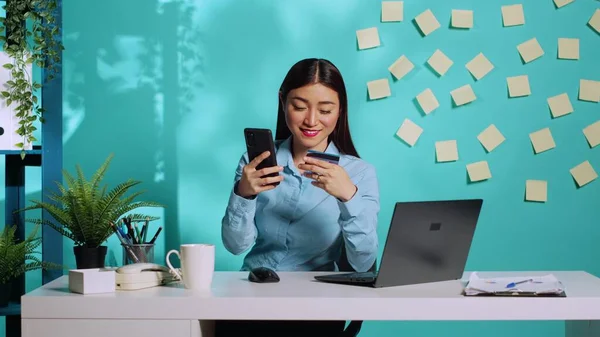 Happy asian employee carefully adding card payment information, excited to do online shopping while at work. Businesswoman in bright colourful modern workplace over blue studio background