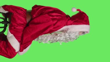 Noel Baba kılığındaki bir adamın dikey video profili yeşil ekran arkaplanına karşı oturan bir sandalyede oturuyor, Noel Baba 'nın vücut bulmuş hali. Genç yetişkin reklamı Aralık mevsimsel tatil, geleneksel etkinlik.