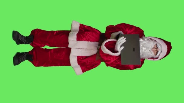 垂直ビデオサンタのフロントビューでは 白いひげとブラウジングウェブサイトでお祝いのクリスマスの衣装を着て 緑の画面上のラップトップを使用しています 現代の聖人ニックは ポータブルPc上で動作します スタジオでのオンラインネットワーク — ストック動画