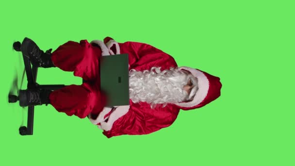 垂直方向のビデオ椅子にラップトップを使用してクールな聖人ニックのフロントビュー スタジオでフルボディの緑の背景に対して座って 父のクリスマスは現代のポータブルPcで作業します オンラインウェブサイトを閲覧 — ストック動画