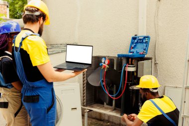 Nitelikli elektrikçi dış klima iç kompresörünü temizleme ve yağlama, freon seviyesini kontrol ederken Afrikalı Amerikalı meslektaşı dizüstü bilgisayardaki bakım programını kontrol ediyor