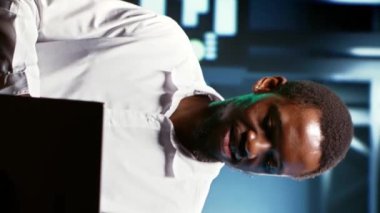 Dikey video Mutlu Afro-Amerikan çalışanı sunucu dolaplarındaki enerji tüketimini incelemek için dizüstü bilgisayar kullanıyor. İşçi, veri merkezinin elektronik sıcaklık sensörlerinin çalıştığından emin oluyor