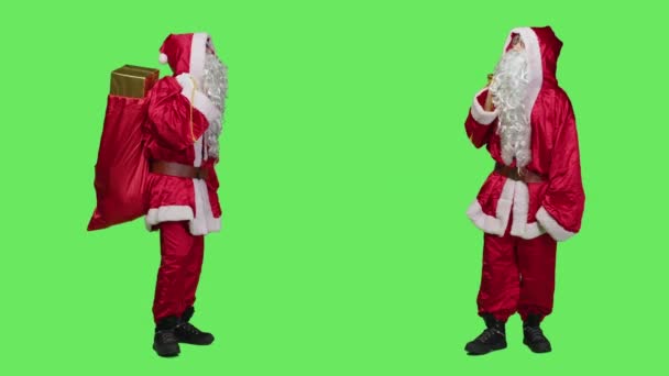緑の背景を持つスタジオでプレゼントの袋を運ぶセントニックは 子供たちにおもちゃを提供する準備をしています 父のクリスマスを描いた若い男の贈り物の袋 休日の精神 — ストック動画