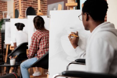 Fiziksel engelli yetişkinler için sanat aktiviteleri. Engelli Afrikalı Amerikalı tekerlekli sandalye kullanıcısı, grup resim dersini ziyaret ederken tuval üzerine kalem çiziyor, terapi seansına katılıyor.