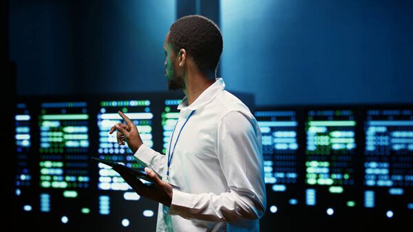 African american supervisor in server farm using tablet to find firewall misconfigurations affecting rackmounts performance and connectivity, leading to slow data transfer rates