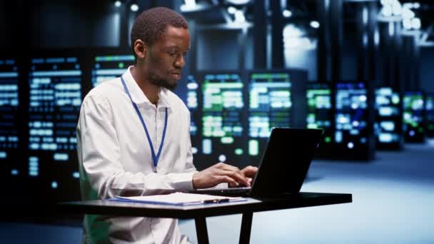 アフリカ系アメリカ人のサービスマンは 企業がデータベースを管理しファイルを保存するためのメインフレームを評価する 高性能コンピューティングに使用される専門的な検査サーバーファームエレクトロニクス — ストック動画