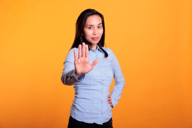 Negatif Asyalı kadın fotoğraf makinesinin önünde el hareketi yapıyor. Stüdyoda sarı arka planda poz veriyor. Ciddi genç yetişkinler jestleri durdurur, reddeder ve reddeder..