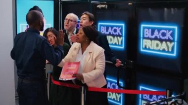 Anxious customers in line for deals on black friday, standing behind red tape at clothing store entrance. Impatient shoppers waiting for opening shop to start promotional shopping spree