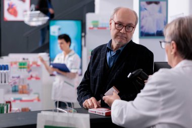 Yaşlı müşteriler eczane kasasında kasiyer arıyor. Ek gıda fiyatlarını kontrol ediyorlar. Yaşlı adam eczaneden alışveriş yapıyor, sağlık vitamini alıyor ve resepsiyonun yanında duruyor.