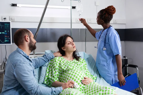アフリカ系アメリカ人看護師は妊娠中の女性の点滴をチェックし 病棟での医療手術の準備をします 妊娠中の患者はベッドに横になり 親について夫と話し合う — ストック写真