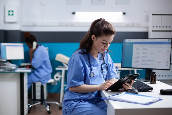在繁忙的医务室 年轻护士在数字平板电脑上查看预约名单 在医院服务台与技术打交道的成年妇女保健专家 脖子周围有听诊器 — 图库照片