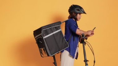 BIPOC bisikletçisinin müşterilere yemek dağıtırken, telefon uygulamasında bir sonraki adresi yazarken elde çekilmiş bir fotoğrafı. Kurye bisikleti, koruyucu giysi ve termal çanta, stüdyo arka planında izole.