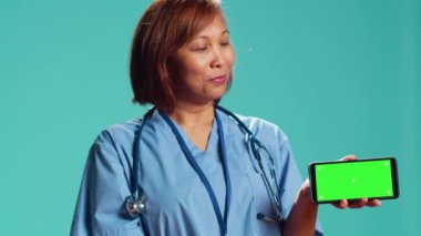 Stüdyonun arka planında izole edilmiş klinik çalışanının yakın çekimleri kroma anahtar telefon yeşil ekranında tıbbi bandı gösteriyor. Hemşire akıllı telefonu peyzaj modunda tutuyor, bilgilendirici video sunuyor