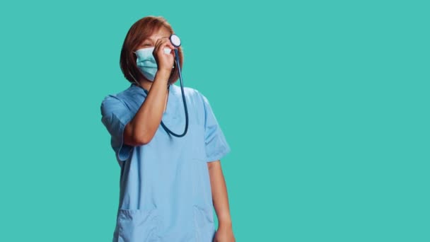 アジアの経験豊富な看護師は プロの聴診器を使用して適切に表示するモックを行います 青いスタジオの背景に隔離された医療機器の使用法を示す知識豊富なクリニックの労働者 — ストック動画