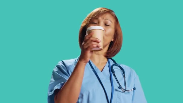 満足しているBipocヘルスケアの専門家は 仕事のシフト休憩中に新鮮なコーヒーを楽しんでいます ハッピー認定看護師 スタジオのバックグラウンドで孤立 ホットドリンクを飲む 閉じる — ストック動画