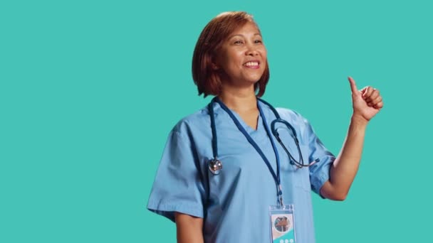 乐观的Bipoc专家护士在工作时竖起大拇指 在工作室背景上被隔离 近距离拍摄快乐的亚洲医疗专家做认可手势 — 图库视频影像