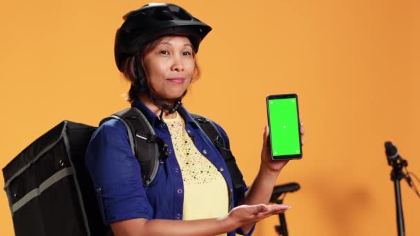 クロマキーグリーンスクリーンスマートフォンをモックアップする女性のクローズアップ スタジオの背景に隔離された顧客の住所にバイサイクルに乗る準備をする安全ヘルメットを身に着けている急使の肖像画 — ストック動画