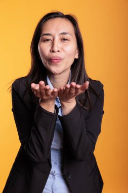 Filipino woman blowing air kissed during studio shot, posing over yellow background. Confident positive young adult expressing love, looking at the camera with a satisfied smile. Romantic gesture