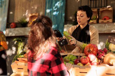 Young female farmer talking with customer while selling fresh locally grown vegetables at farmers market, local vendor helping consumer to choose veggies, explaining health benefits of organic food