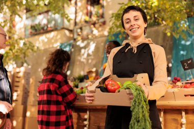 Young cheerful seller fruits and veggies market stand owner showing fresh organic produce box and smiling at camera. Female farmer selling healthy local food from roadside stand, bio products.
