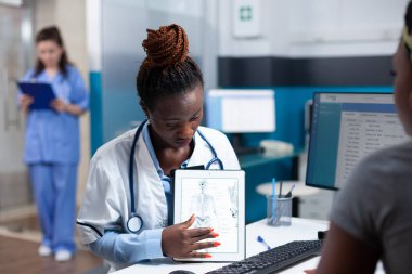 Bone doctor explaining clinical information on tablet to patient during orthopedic appointment. Orthopedist in modern hospital office and african american woman at medical checkup