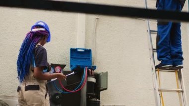 Precise worker measuring refrigerant levels in air conditioner with professional manometers while coworker steps down from folding ladder after finishing checking rooftop HVAC system