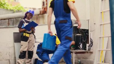 Expert african american technician cleaning layer of dirt and dust from condenser compressor coils while colleague uses manometers to check for refrigerant leaks. Read pressure in hvac system.