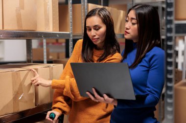 Depo teslimat müdürleri perakende satış deposunda dizüstü bilgisayarda sipariş işleme sistemi kullanıyorlar. Asyalı genç kadın çalışanlar raflardaki karton kutuları arıyorlar.
