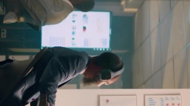 Dikey video Afrikalı Amerikalı iş adamı bilgisayardaki istatistiksel grafikleri kontrol ederken müzikten zevk alıyor. Gece vardiyası sırasında ofisteki proje görevlerini çözmek için kulaklıkları olan rahat bir çalışan.