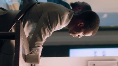 Gece vardiyasında masa başında oturan Afrikalı Amerikalı bilgisayar operatörünün dikey video portresi arka plandaki iş arkadaşı tuvaletteyken internetteki finansal iş belgelerine bakıyor.