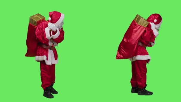 有名な赤いバッグの贈り物でクリスマスの精神をスタジオで広めるお祝いのコスチュームで聖なるニック サンタクロースは フルボディグリーンを見渡すプレゼントの袋を体現しています — ストック動画