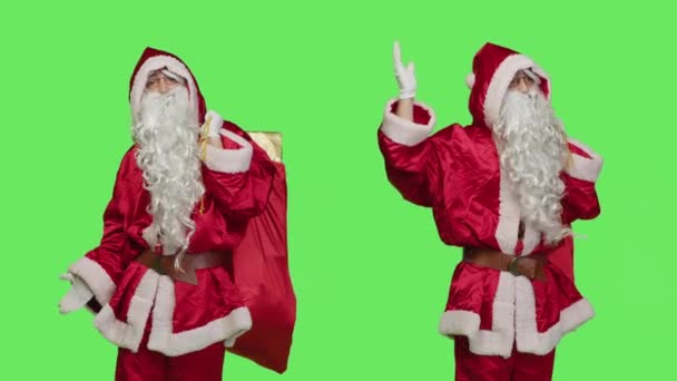 サンタのキャラクターは 彼が贈り物を持ってバッグを運んで 有名な衣装を着ている間 何かを脇に示しています 緑色の背景に聖なるニックとして偽装した男 — ストック動画