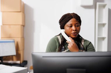 Depo çalışanı sabit hatta uzaktan müşteriyle konuşuyor. Depoda teslimat detaylarını tartışıyorlar. Depo amiri depodaki bilgisayardan online siparişleri kontrol ediyor