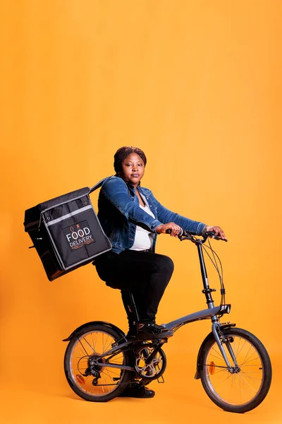 Restaurant worker riding bike while carrying takeaway thermal backpack ready to deliver order to customer during lunch time, standing over yellow background. Take out concept and transportation