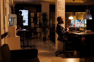 Uzaktan kumandalı ekiple çevrimiçi görüşme yapan bir iş adamı, video görüşmesi sırasında pazarlama istatistiklerini tartışıyor. İş stratejisinde fazla mesai yapan çeşitli meslektaşlar