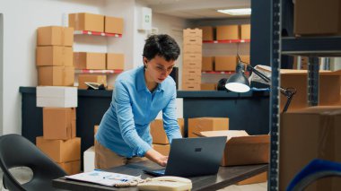 Caucasian woman putting merchandise in packages, working with laptop to do stock inventory before shipping products to customers. Female worker planning delivery management and logistics.