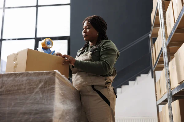 工人使用胶带包装客户的订单 准备产品在存储室装运 非洲裔美国人雇员穿着工业整体工作服在仓库的货物库存处工作 — 图库照片