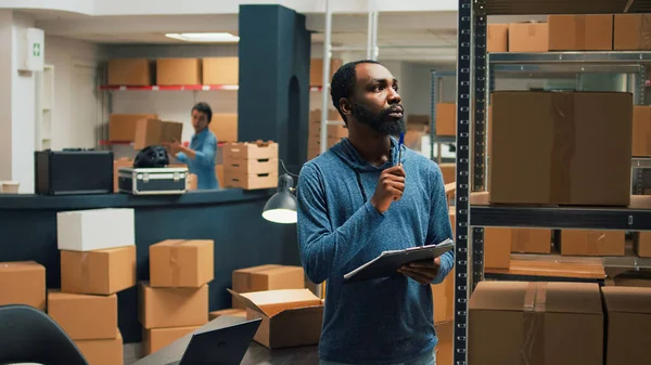 African american employee taking inventory notes on clipboard papers, checking quality of products and packages. Young adult looking at cardboard boxes, merchandise stock logistics.
