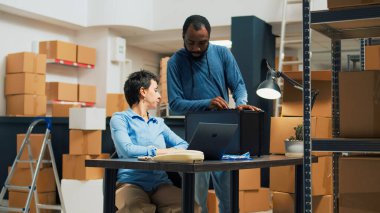 Entrepreneurs team working on stock shipping with laptop, preparing order in cardboard package. Man and woman putting products in boxes, doing quality control before distribution. Tripod shot.