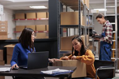 Tekerlekli sandalyedeki sipariş toplayıcısıyla birlikte çalışırken depo teslimat müdürü paket kalitesini kontrol ediyor. Depo sevkiyatı operatörleri dağıtım için karton kutu hazırlıyorlar.