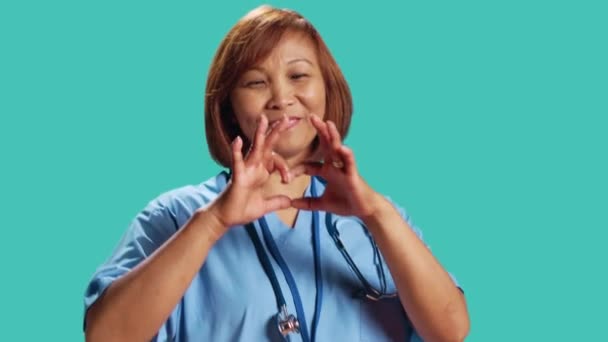 ハートシンボルを手で形作る喜びに満ちたBipoc看護師のクローズアップショット 仕事中に愛のジェスチャーを示すクリニックの従業員は ブルースタジオの背景に孤立しました — ストック動画