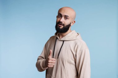 Arap adam başparmağıyla onaylıyor ve kameraya bakıyor. Gündelik kapüşonlu kıyafetler giyen genç adam, anlaşma imzalı stüdyo resminden memnun olduğunu ifade ediyor.