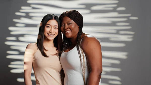 Flawless confident diverse girls posing together on camera, showing self confidence and body positivity. Two beautiful skincare models hugging and promoting different bodies and skintones.