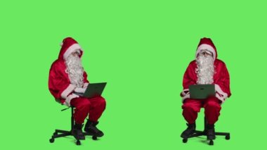 Aziz Nick 'in sandalyede dizüstü bilgisayarı kullanması, stüdyoda tüm yeşil ekran arka planına karşı oturması. Noel Baba, modern taşınabilir bilgisayarla çalışıyor. Kırmızı kostümlü internet sitesine göz atıyor..