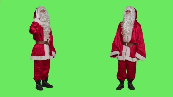 フレンドリーなキャラクターの波とカメラで人々を迎え クリスマスの精神を広めるために伝統的なお祝いの衣装を着ます サンタクロースの格好をした若い大人 こんにちは 緑色の背景 — ストック動画